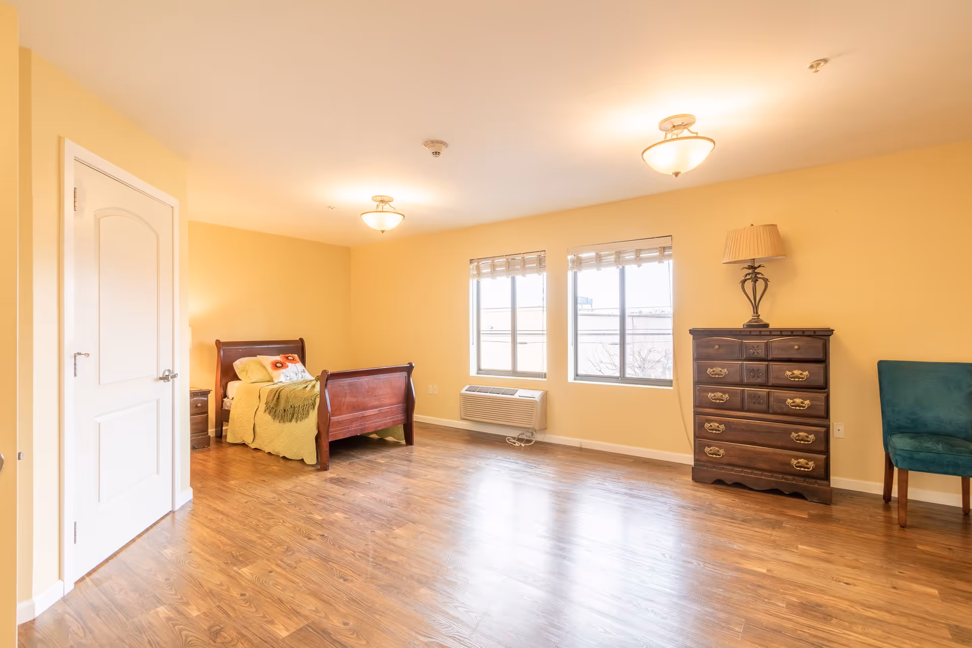 A bright bedroom with yellow walls and wooden flooring. The room features a wooden bed with green bedding and a decorative pillow, a wooden dresser with a lamp on top, a teal upholstered chair, two windows with blinds, and a white door on the left side.