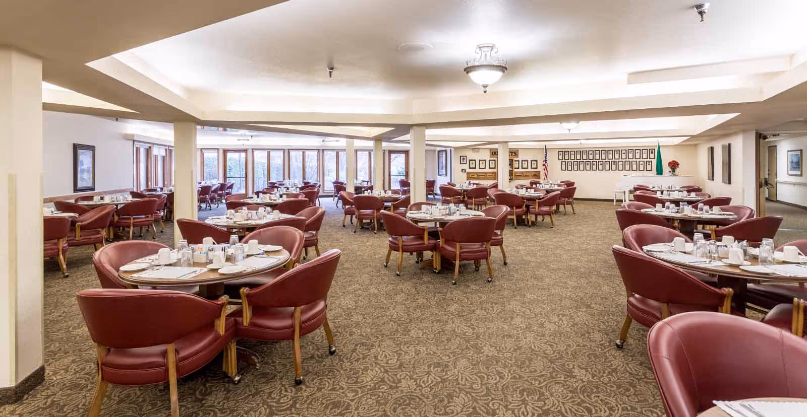 Spacious dining room with multiple round tables set with white cups, glasses, and silverware. Each table is surrounded by red cushioned chairs with wooden legs. The room has beige walls, patterned carpet, and large windows letting in natural light. There are framed pictures on the walls and a white piano in the back corner.