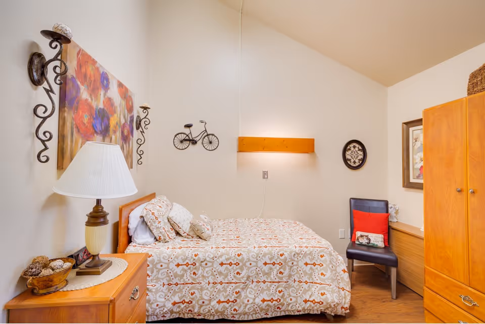 A cozy bedroom with a bed covered in a patterned bedspread and multiple pillows. There is a wooden nightstand with a lamp and decorative items on it. The walls are decorated with a colorful floral painting, a small metal bicycle wall art, and a round clock. A black chair with a red pillow and a wooden wardrobe are also visible in the room.