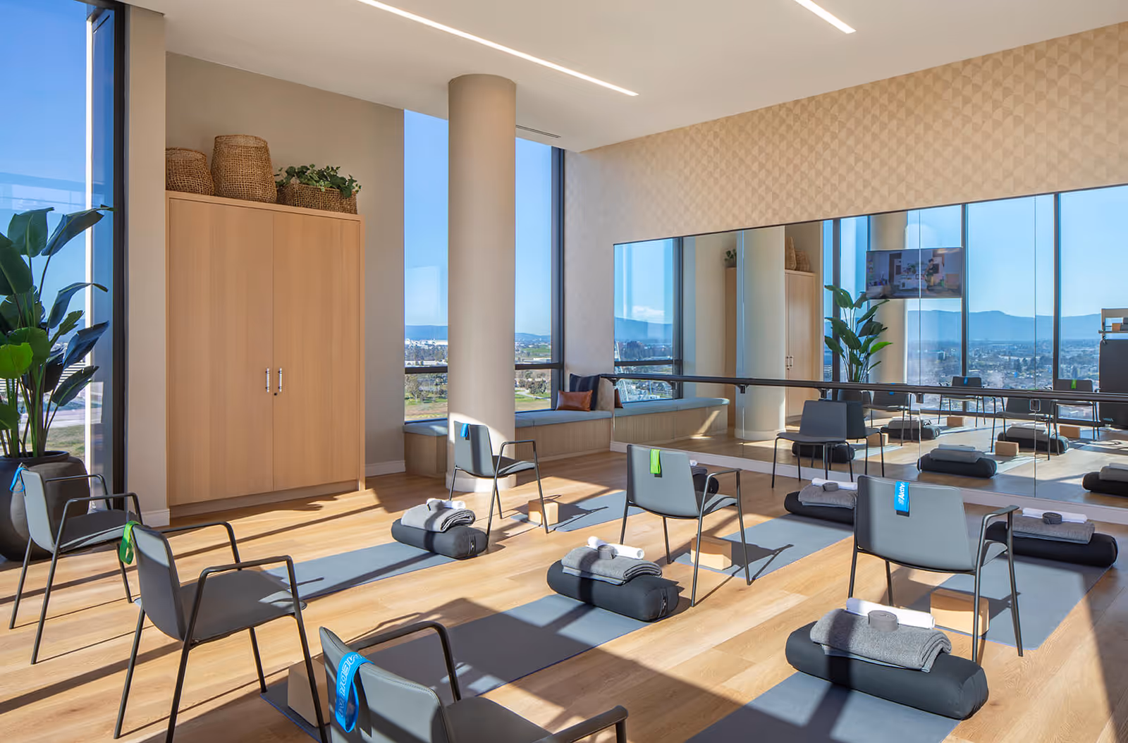 Bright and spacious exercise or yoga room with large windows offering a scenic view. The room has wooden floors, several chairs placed on yoga mats with rolled towels and blocks, a large mirror on one wall, a wooden cabinet with baskets and plants on top, and a potted plant near the window.