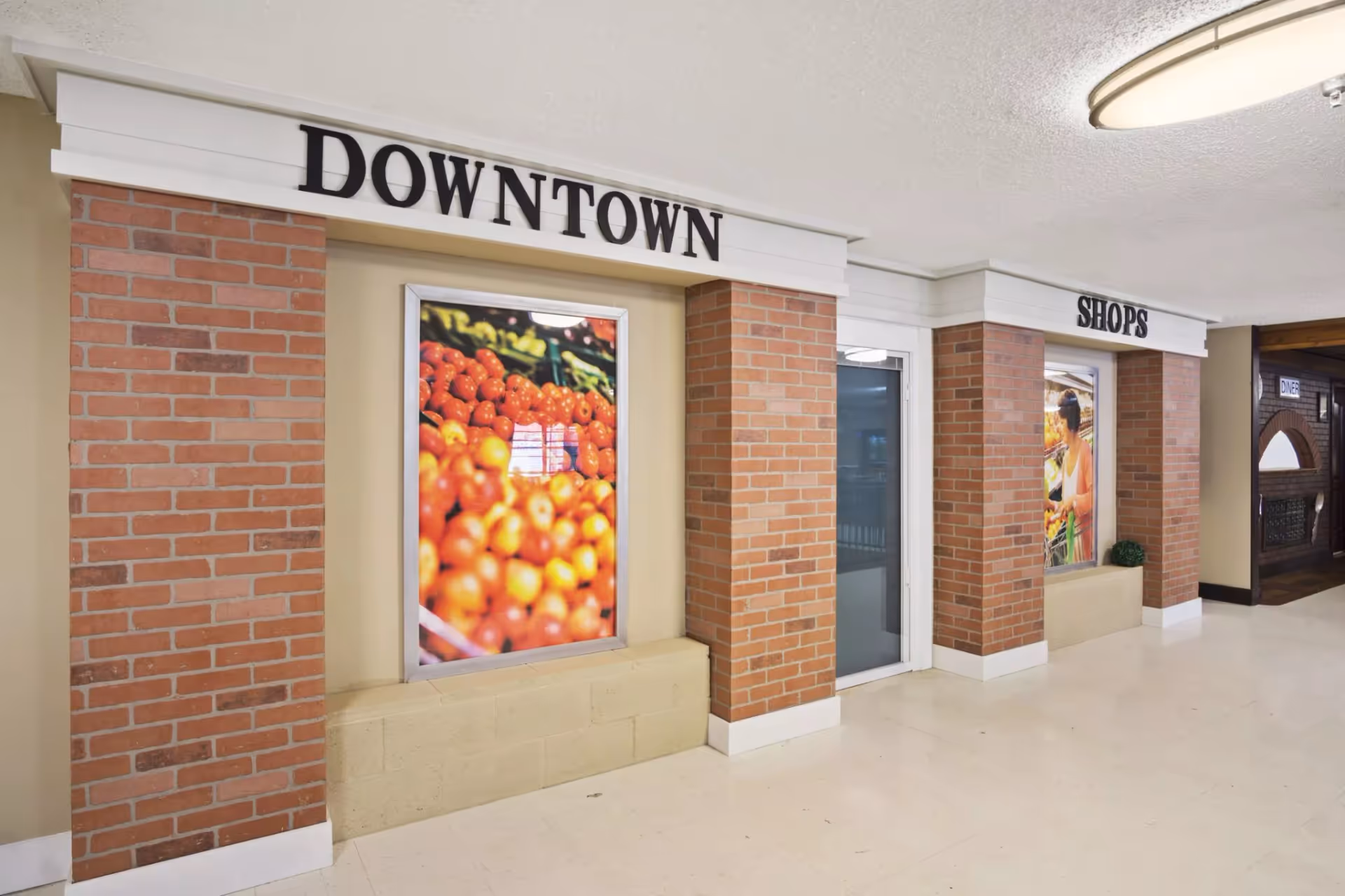 Interior hallway styled as a small downtown shopping area with brick columns, storefront signs reading "DOWNTOWN" and "SHOPS", framed produce posters and a glass door.