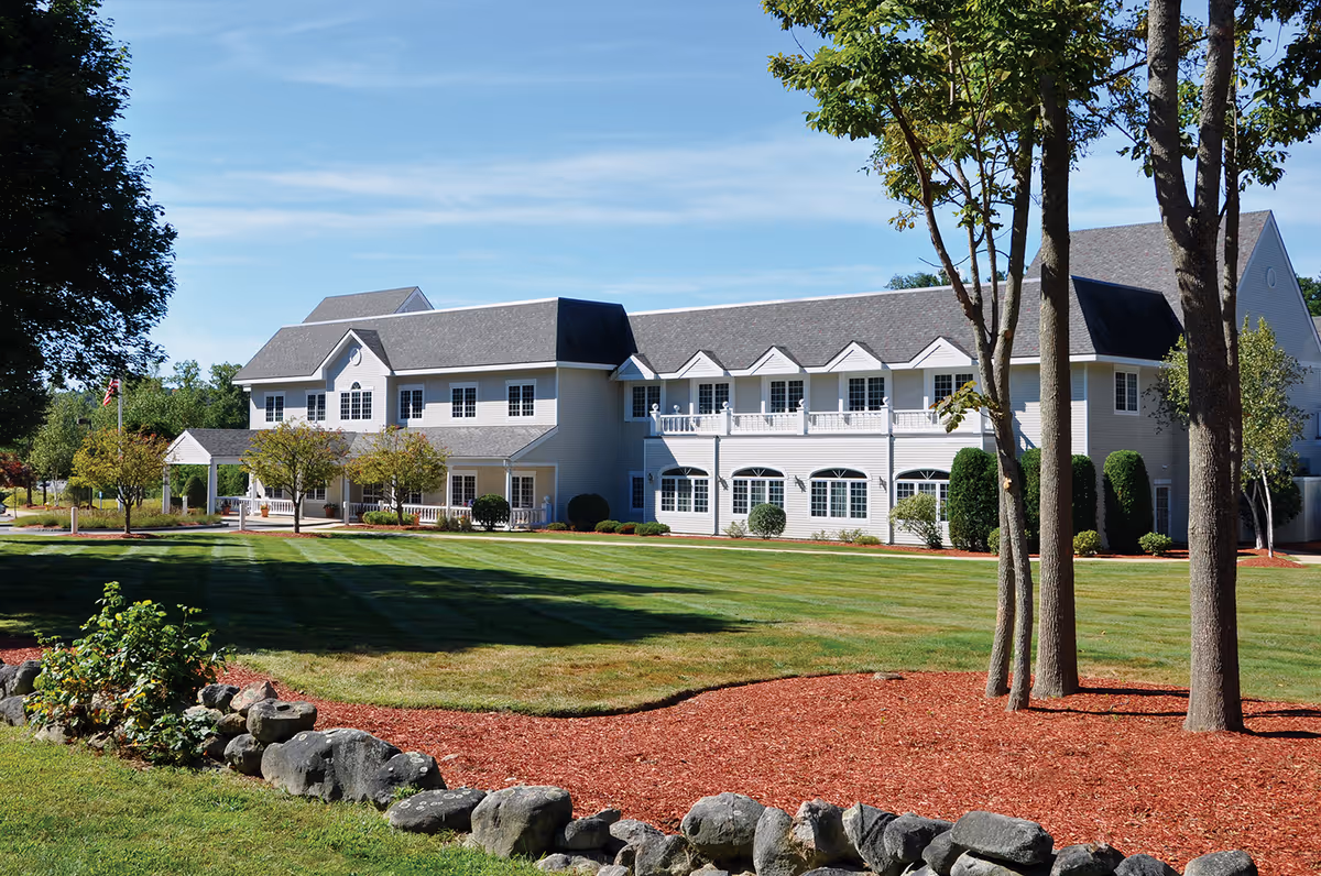 A large two-story healthcare center building with a gray roof and white exterior surrounded by a well-maintained lawn, trees, and landscaped areas with rocks and mulch under a clear blue sky.