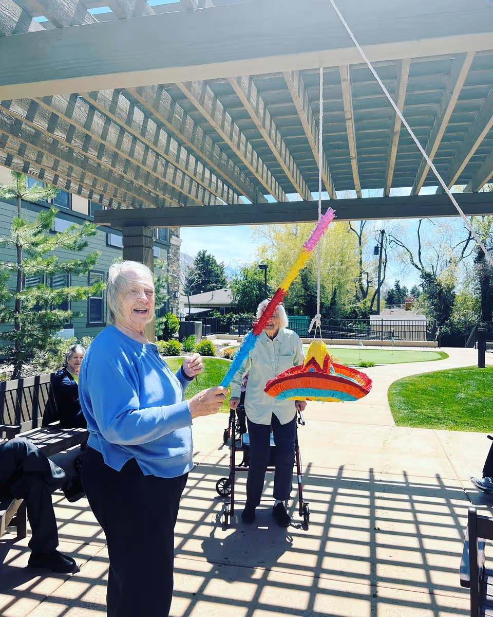 Two elderly women outdoors under a wooden pergola at a senior living facility. One woman in a blue sweater is holding a colorful stick and smiling, preparing to hit a sombrero-shaped piñata hanging from the pergola. Another woman with a walker stands nearby, watching. There are benches and greenery around, with a clear sky and trees in the background.
