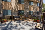 Outdoor patio area with several wooden benches and potted plants, adjacent to a building with multiple windows and a stone and beige exterior.