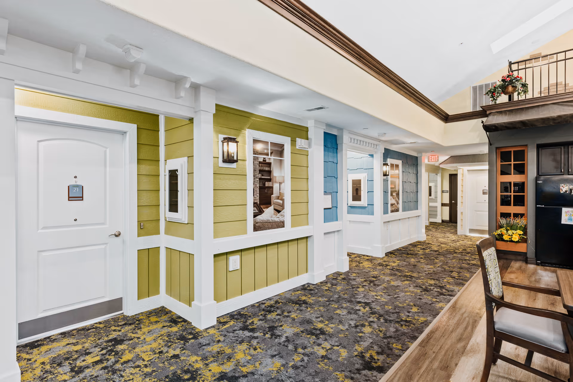 Bright interior corridor styled like a neighborhood street with paneled apartment doors, decorative siding, lanterns, patterned carpet, and a seating area.