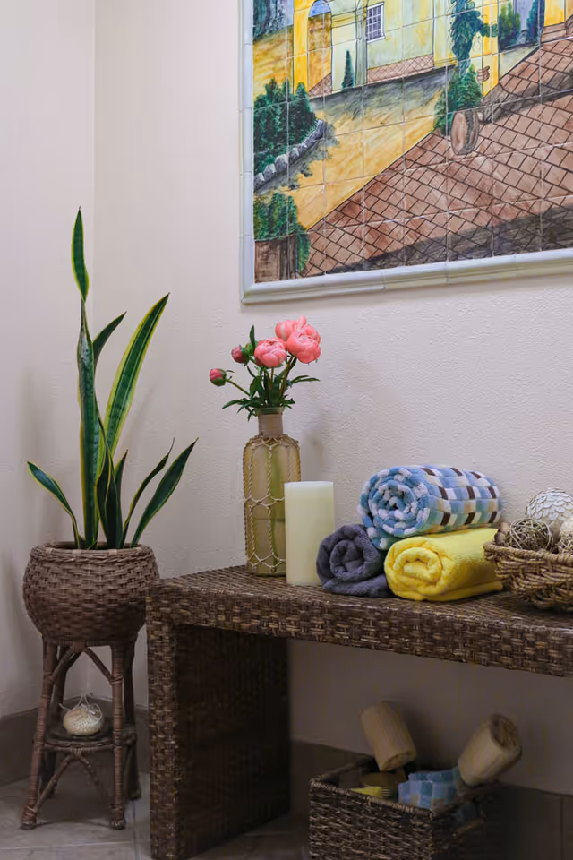 A cozy corner with a woven wicker table holding rolled towels in yellow, gray, and checkered patterns, a beige candle, and a vase with pink flowers. Next to the table is a tall potted snake plant in a wicker stand. Above the table, a framed tile mural depicts a scenic outdoor pathway with greenery and buildings.