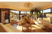 A bright and spacious living room with large windows allowing natural light to fill the space. The room features cream-colored sofas, a wooden coffee table with a floral arrangement, side tables with lamps, a chandelier, and several potted plants. Outside the windows, a patio and garden area are visible.