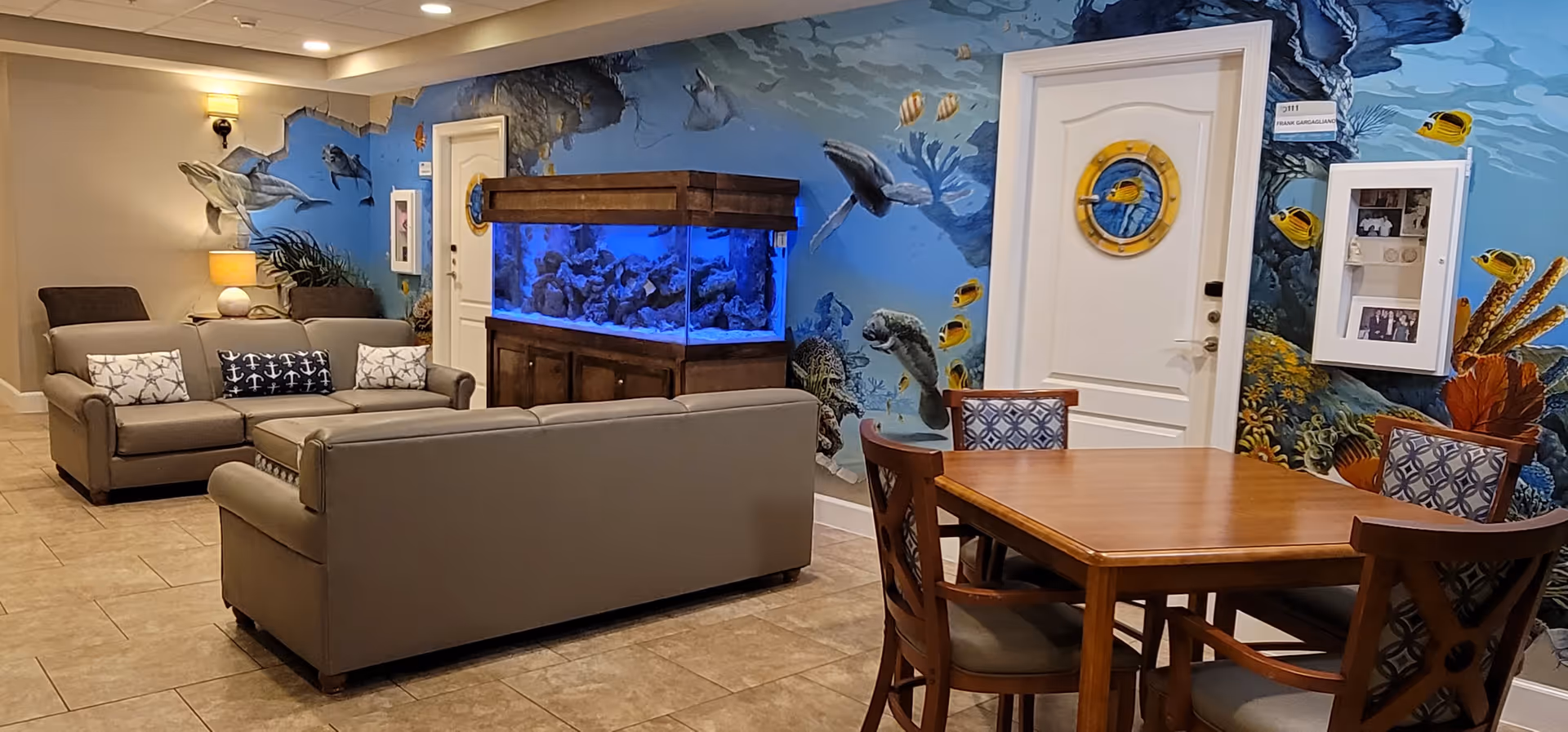 A cozy living room area with two gray sofas, a wooden table with four chairs, and a large aquarium filled with fish. The walls are decorated with a vibrant underwater mural featuring sea turtles, fish, and coral. There is a white door with a nautical-themed porthole window and a wall-mounted display case with photos.