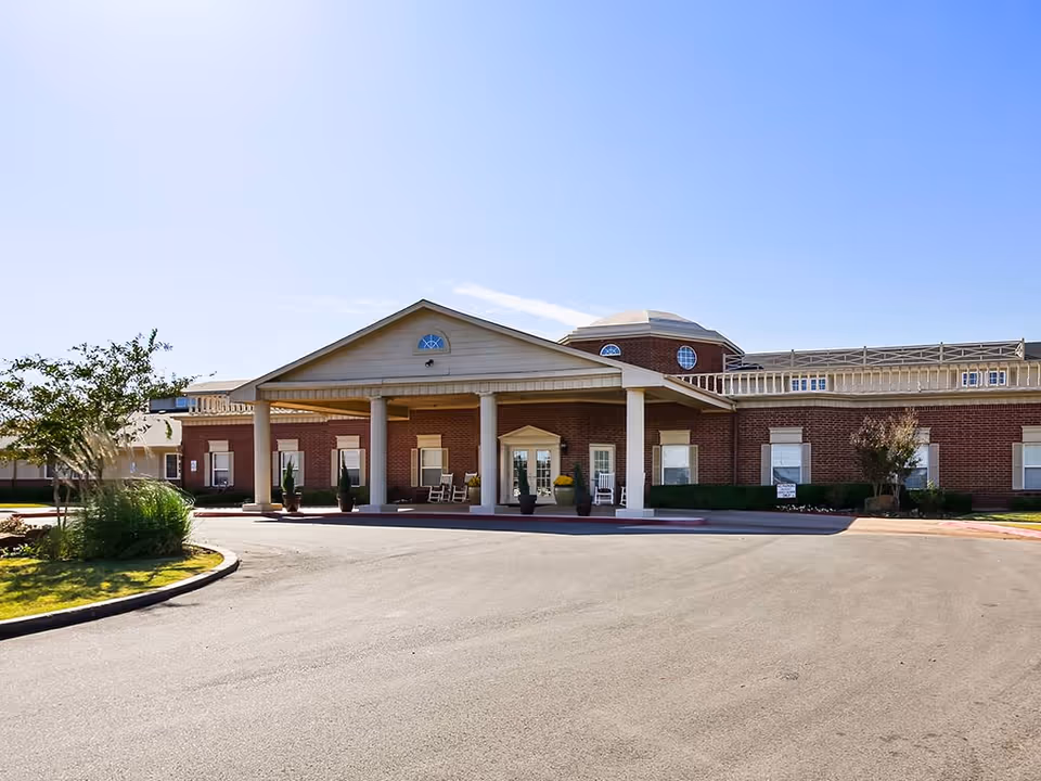 Front exterior view of a single-story brick building with a covered entrance supported by white columns, potted plants, and rocking chairs near the entrance. The sky is clear and blue.
