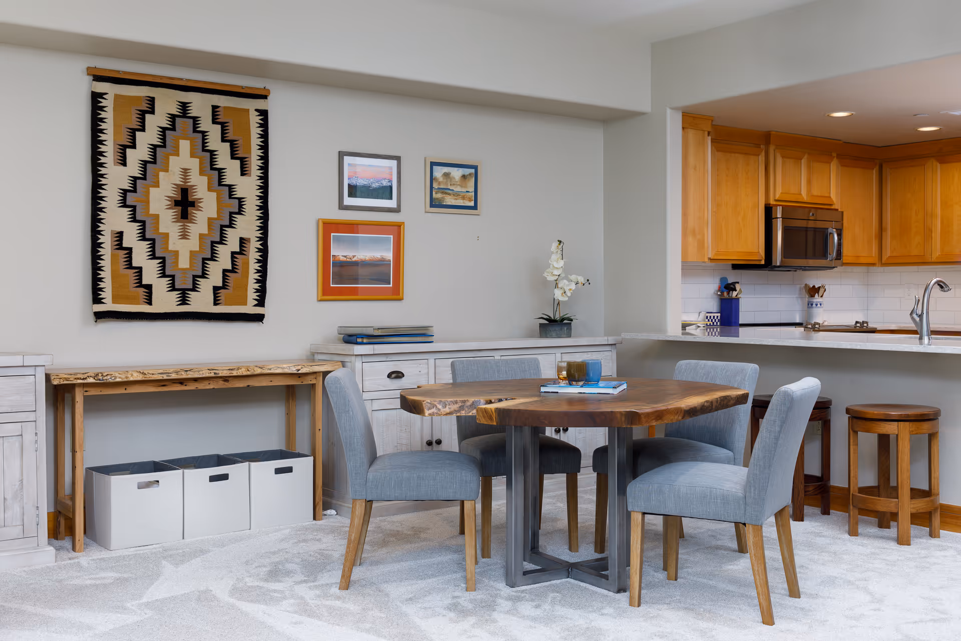 Open dining area with a live-edge wooden table and four gray upholstered chairs, wall art on a light wall and a view into a kitchen with wooden cabinets.