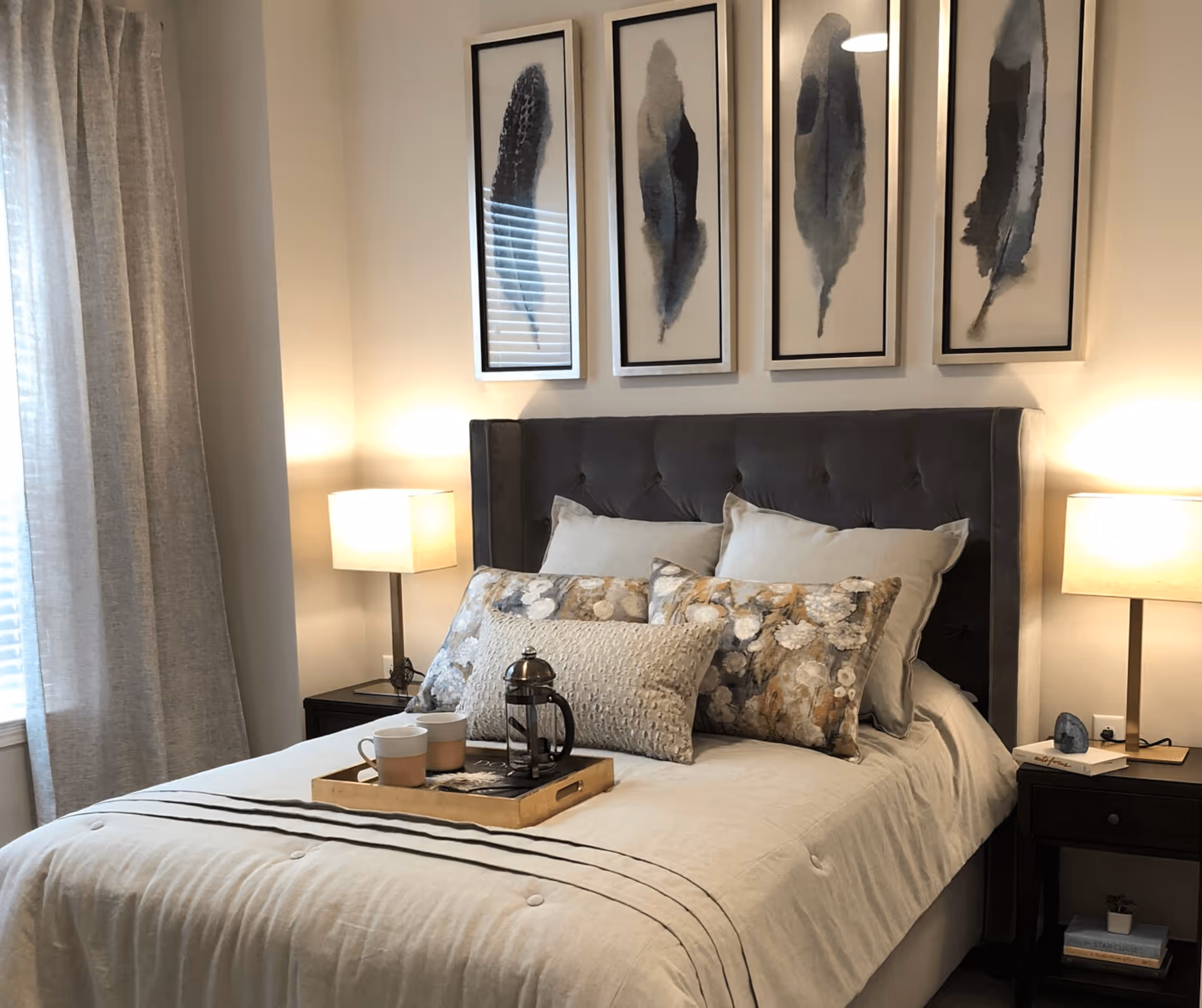A neatly made bed with a tufted dark gray headboard, adorned with multiple decorative pillows in neutral and floral patterns. A wooden tray with two cups and a French press sits on the bed. On either side of the bed are dark wooden nightstands with modern lamps emitting warm light. Above the bed are four framed artworks featuring abstract feather designs. Light-colored curtains cover a window to the left.