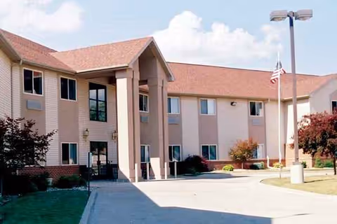 Two-story senior living facility exterior with entrance canopy and American flag on a sunny day.