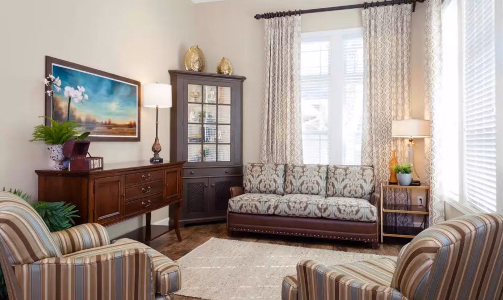Well-lit living room with a patterned sofa, two striped armchairs, wooden sideboard and cabinet, lamps, and decorative accents.