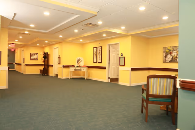 A wide hallway in a senior living facility with yellow walls, green carpet, and ceiling lights. There are framed pictures on the walls, a small table with decorative items, a wooden chair with striped upholstery, and a coat rack in the distance.