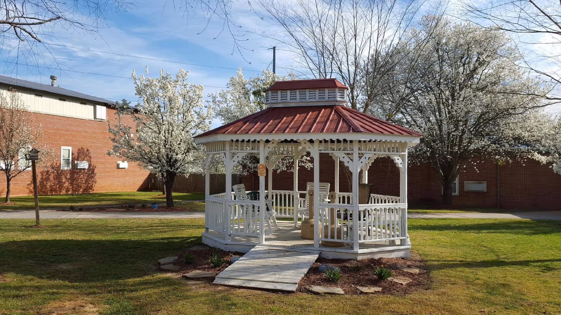 A white wooden gazebo with a red roof situated on a grassy lawn surrounded by blooming white-flowered trees. There are several white rocking chairs inside the gazebo. In the background, there is a red brick building and a clear blue sky.