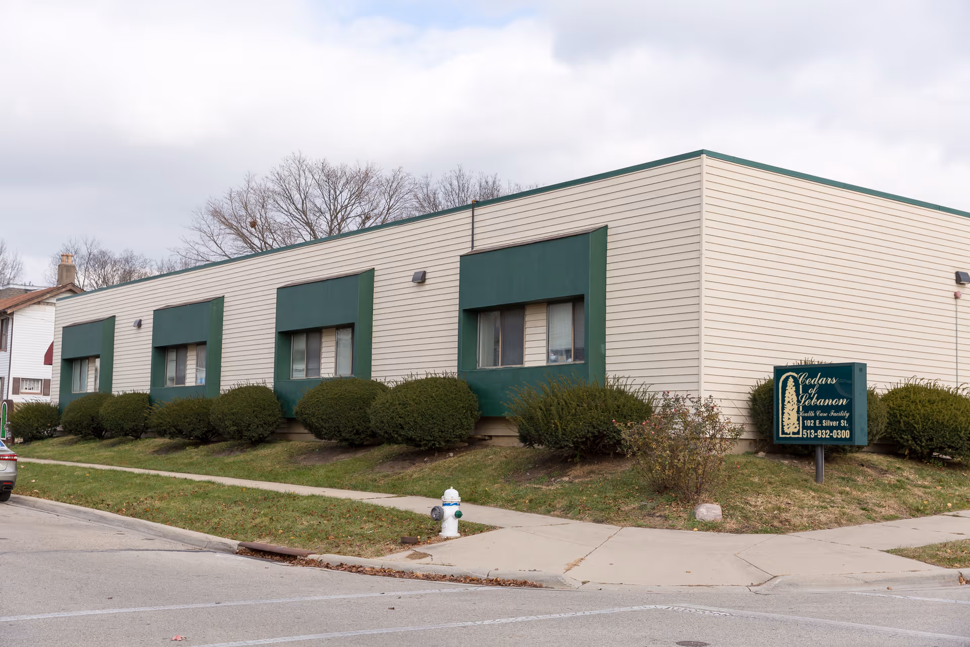 Exterior view of a single-story beige building with green window accents and bushes along the side. A green sign in front reads 'Cedars of Lebanon Rehabilitation & Nursing Care' with the address and phone number. The sky is partly cloudy.