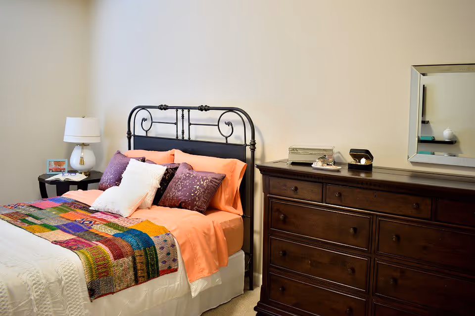Neatly made bedroom with a black metal headboard bed, colorful patchwork throw, bedside table with lamp, and a dark wooden dresser with mirror.