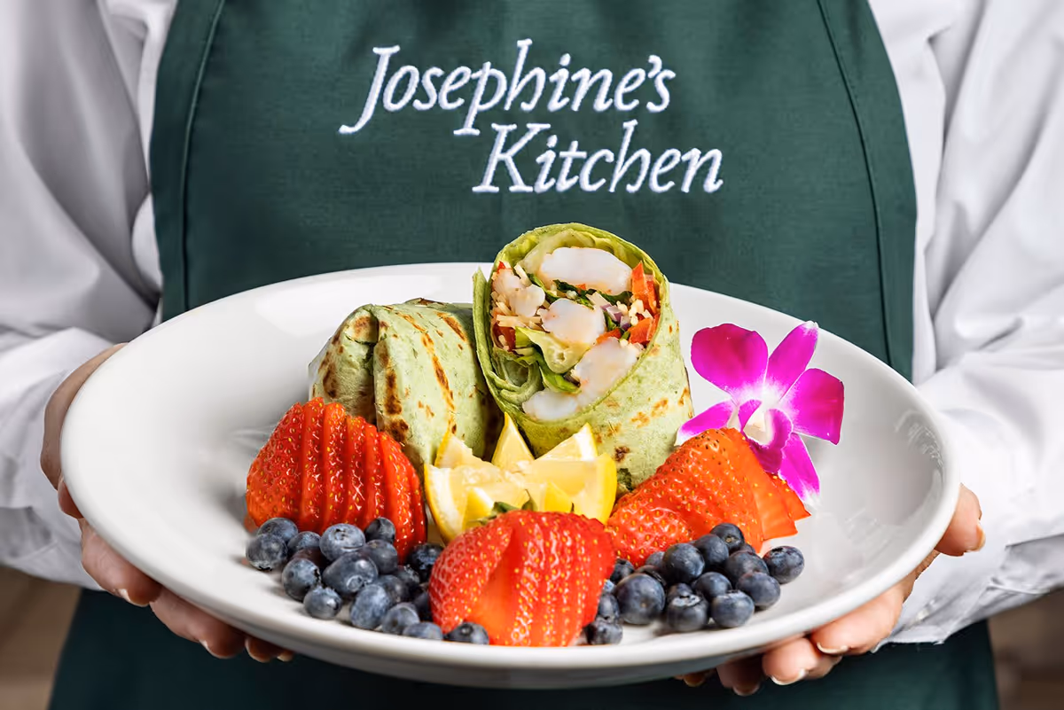 A person wearing a green apron with the text 'Josephine's Kitchen' holds a white plate with a wrap sandwich, sliced strawberries, blueberries, lemon wedges, and a purple orchid flower as garnish.