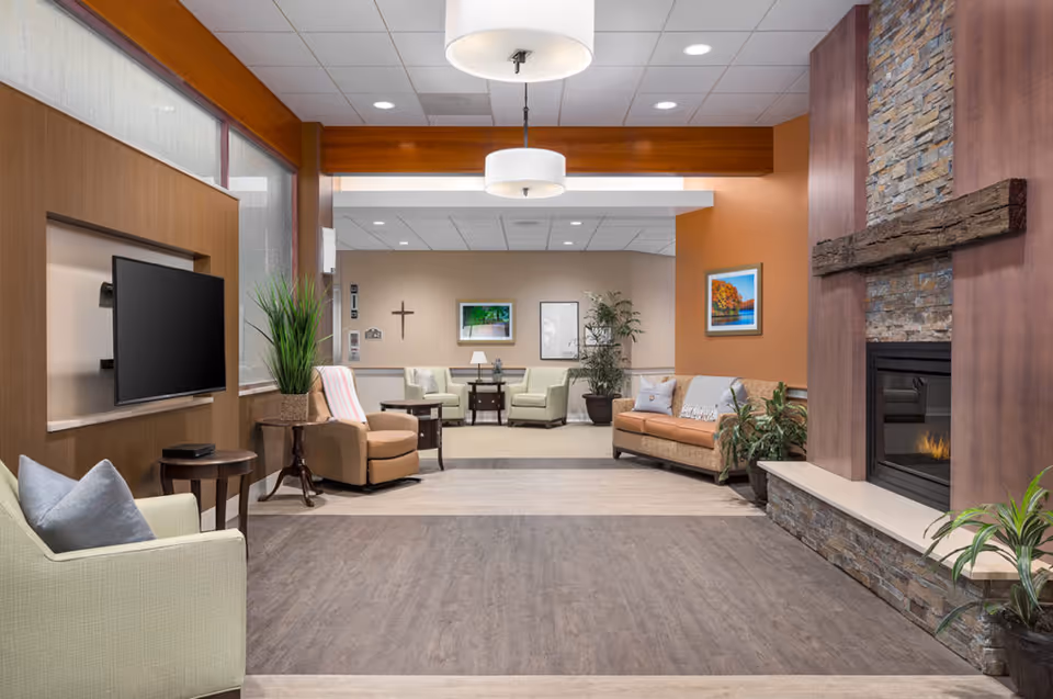 A cozy senior living facility common area with comfortable seating including armchairs and a sofa, a flat-screen TV mounted on a wooden wall, a stone fireplace with a wooden mantel, potted plants, and framed artwork on the walls. The space is well-lit with ceiling lights and has a warm, inviting atmosphere.
