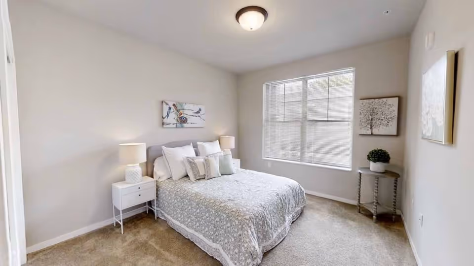 A bright bedroom with a large window covered by blinds. The room features a bed with a patterned bedspread and multiple pillows, flanked by two white nightstands each with a lamp. There are three pieces of wall art, including a bird painting above the bed and two framed pictures on the adjacent walls. A small round table with a potted plant is positioned near the window.