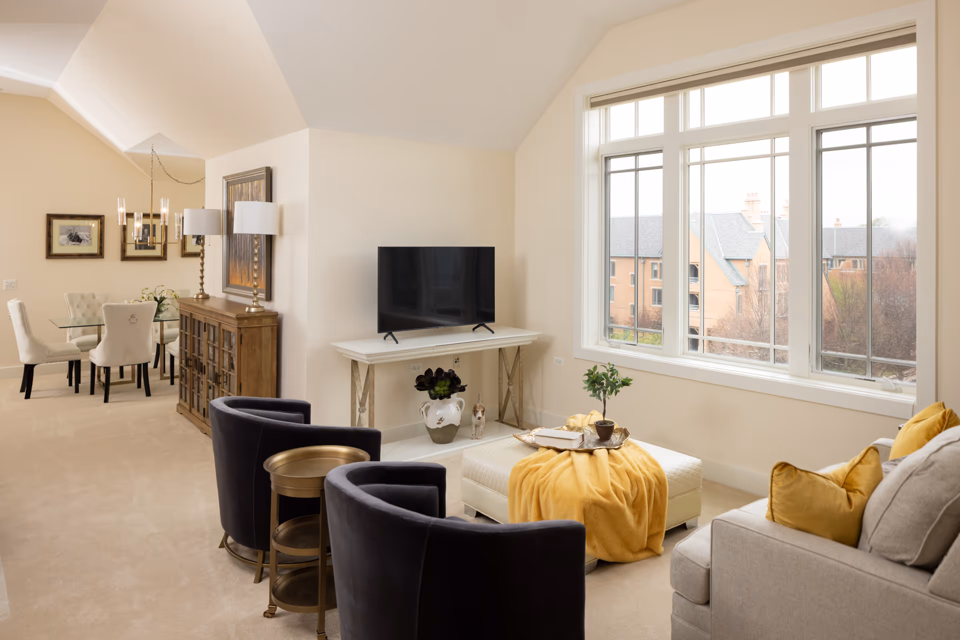 A bright and cozy living room area with large windows showing an outside view of brick buildings. The room features a beige sofa with yellow pillows, two dark gray armchairs, a white ottoman with a yellow throw blanket and a small plant on a tray. A flat-screen TV is placed on a white console table, and a wooden cabinet with two lamps and framed artwork is visible near a glass dining table with white upholstered chairs in the background.