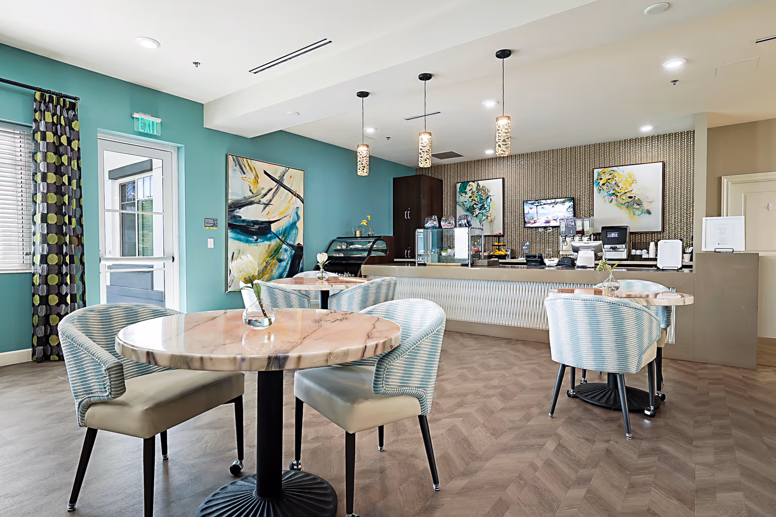 Bright dining area with round marble-top tables, upholstered chairs, and a service counter/coffee bar against a teal accent wall.