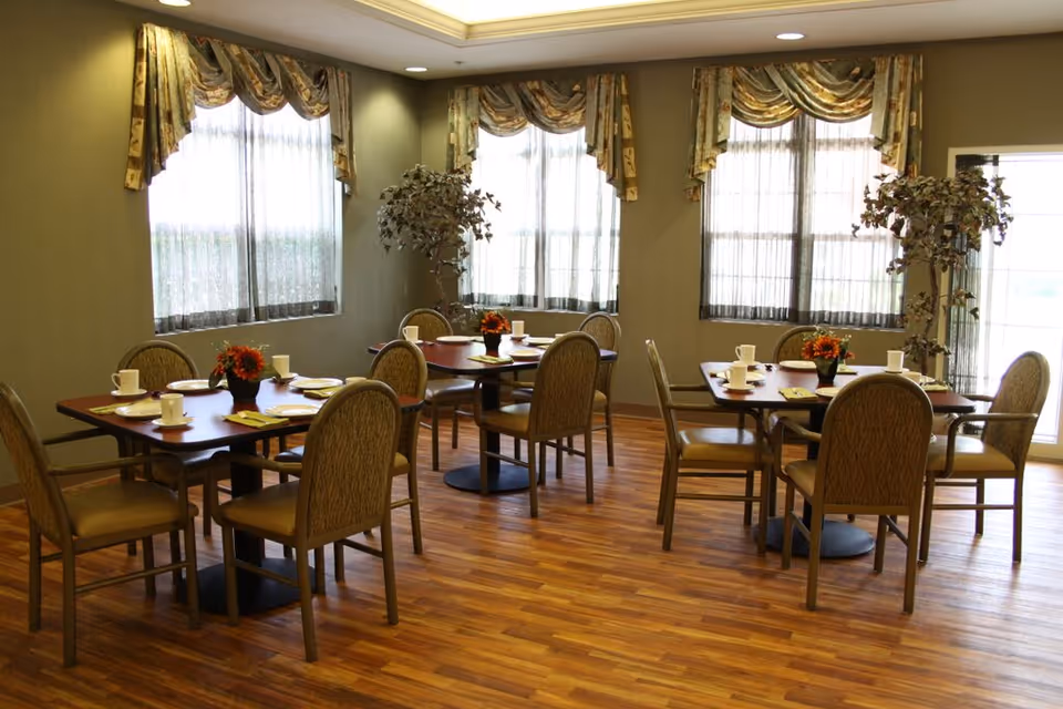A dining room with wooden floors and several tables set with white cups, plates, and napkins. Each table has four chairs with cushioned seats and backs. The room has large windows with sheer curtains and decorative valances, and there are two artificial plants in the corners.