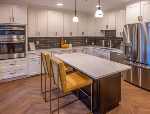 Modern kitchen with white cabinetry, stainless steel appliances including a double oven and refrigerator, a large island with a white countertop, and two yellow cushioned bar stools. The backsplash is dark gray tile, and there are pendant lights hanging above the island.