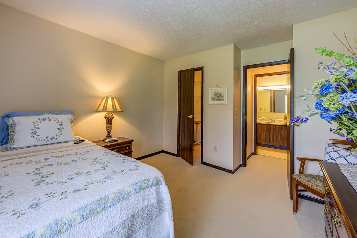 A bedroom with a bed covered in a white quilt with floral patterns, a wooden nightstand with a lamp, an open door leading to a bathroom with a sink and mirror, and a wooden chair next to a table with a vase of blue and green flowers.