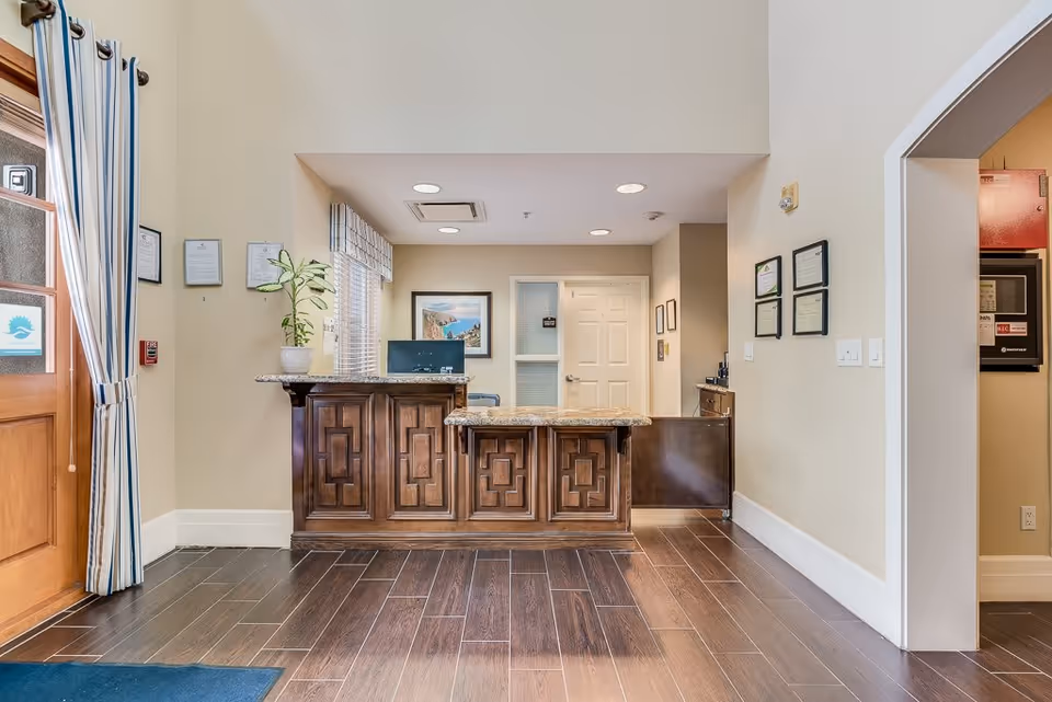 Reception area of a senior living facility with a wooden front desk featuring a granite countertop. Behind the desk is a computer monitor, a potted plant, framed pictures on the walls, and a door. The floor is dark wood, and there is a wooden door with a window and striped curtains on the left side.