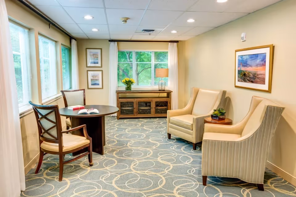 Bright sitting area with two upholstered armchairs, a round table with chairs, a cabinet under a window, and framed artwork.