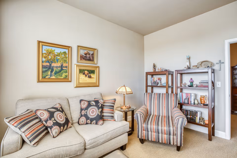 A cozy living room area with a beige sofa adorned with patterned cushions, a striped armchair, a small round side table with a decorative lamp, two wooden bookshelves filled with books and decorative items, and three framed paintings on the wall above the sofa.