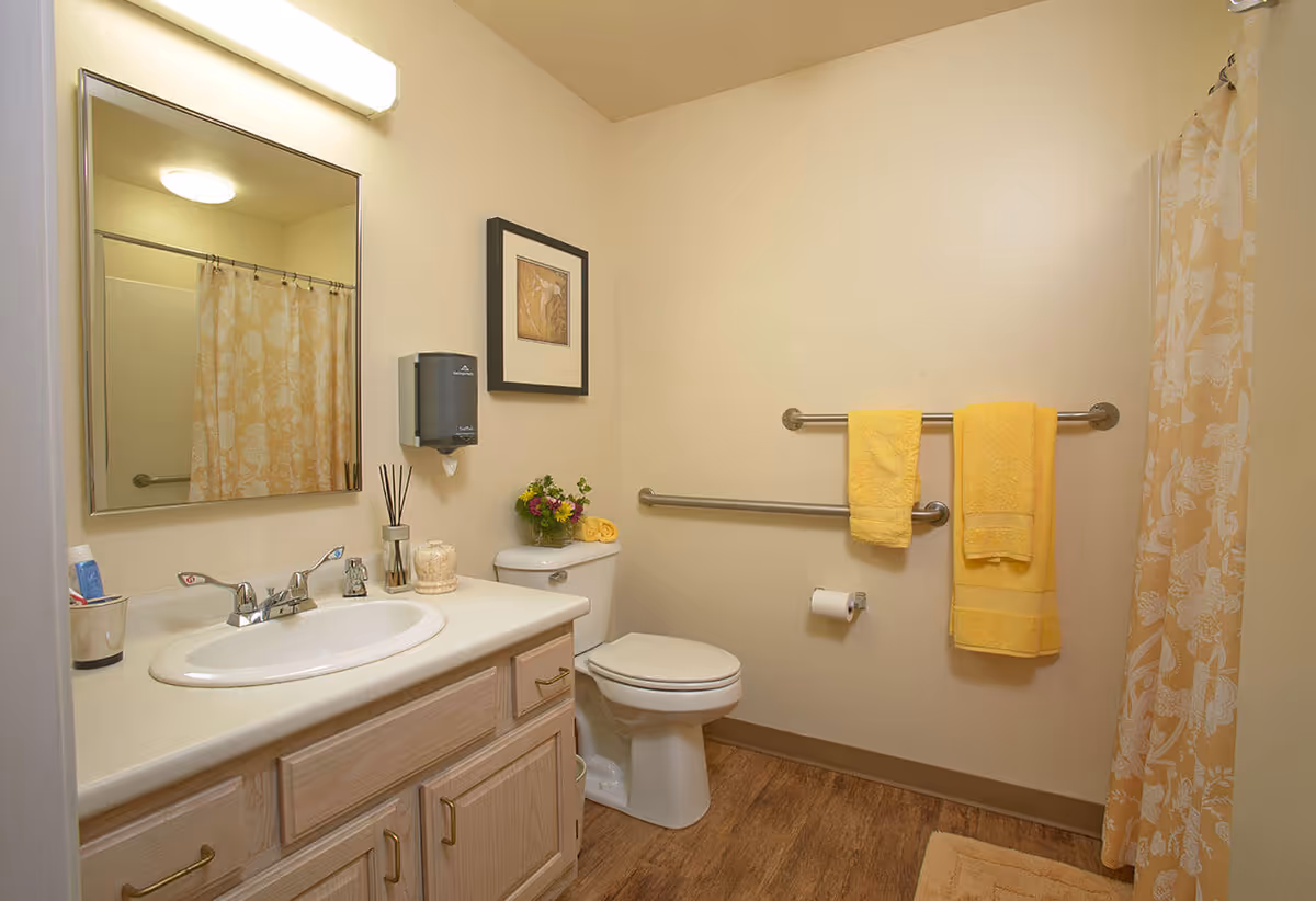 A clean bathroom with a white sink and countertop, wooden cabinets, a large mirror above the sink, a toilet with a small flower arrangement on top, yellow towels hanging on metal grab bars, and a shower with a yellow floral curtain.