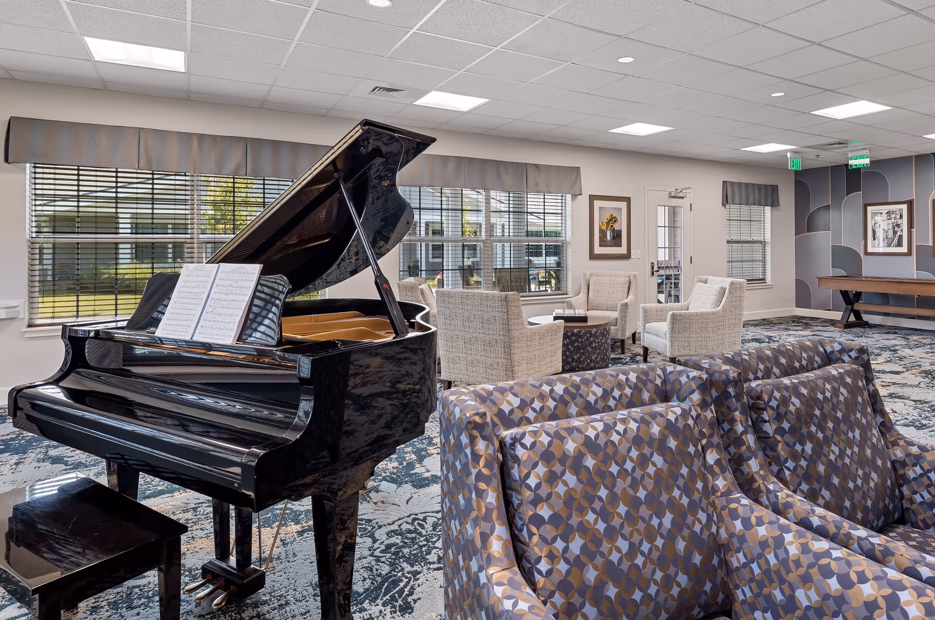 A spacious, well-lit lounge area featuring a black grand piano with sheet music on the stand. The room has patterned blue and beige armchairs, a small seating area with beige upholstered chairs around a round table, large windows with blinds, and modern wall art. The ceiling has recessed lighting and there is a door leading outside.
