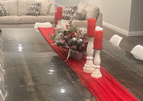 A large wooden dining table with a red table runner, decorated with a centerpiece of greenery and red berries, and three white candle holders with red candles. In the background, there is a beige sofa with patterned cushions in a living area.