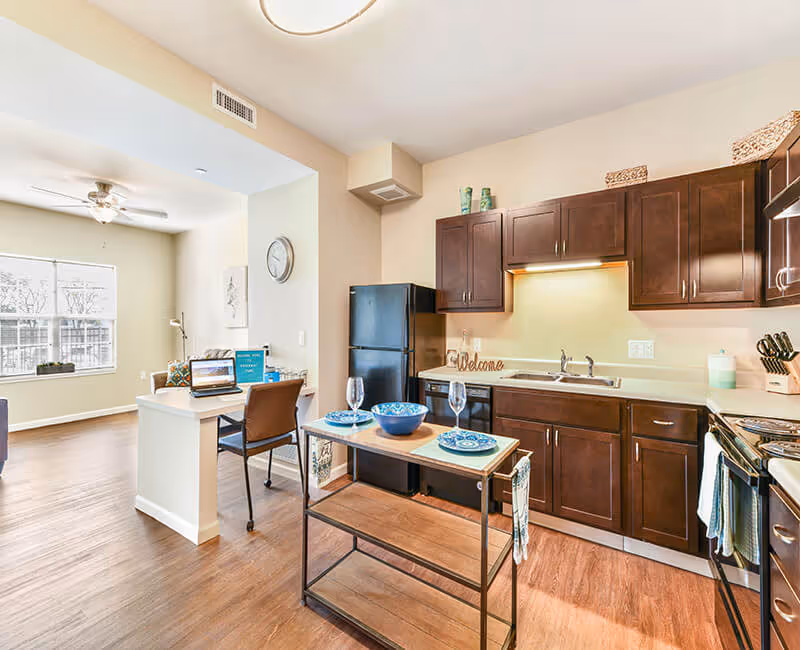 Bright and modern kitchen area with dark wooden cabinets, a black refrigerator, a stove, and a small island table set with blue plates and glasses. Adjacent to the kitchen is a small workspace with a chair and a laptop, and a large window letting in natural light in the background.