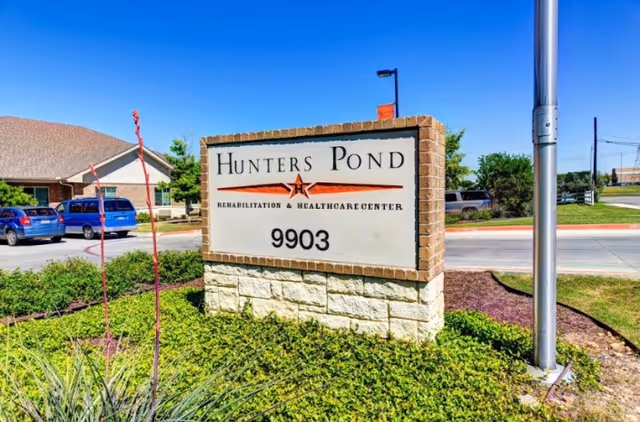 Outdoor view of a sign for Hunters Pond Rehabilitation and Healthcare Center with the address number 9903. The sign is made of brick and stone and is surrounded by greenery. In the background, there is a building, parked cars, and a clear blue sky.
