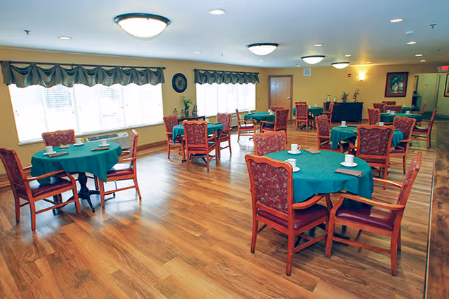 Dining room with round tables covered in green tablecloths and wooden chairs on a wood floor beneath ceiling lights.