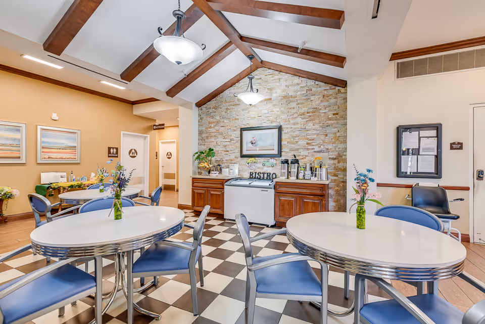 A bright and clean dining area in a senior living facility with white oval tables and blue chairs. Each table has a small vase with flowers. The room features a checkered black and white floor, a stone accent wall with a framed picture, and a buffet area labeled 'Bistro' with coffee dispensers. There are two restroom doors visible in the background and decorative ceiling beams with hanging lights.