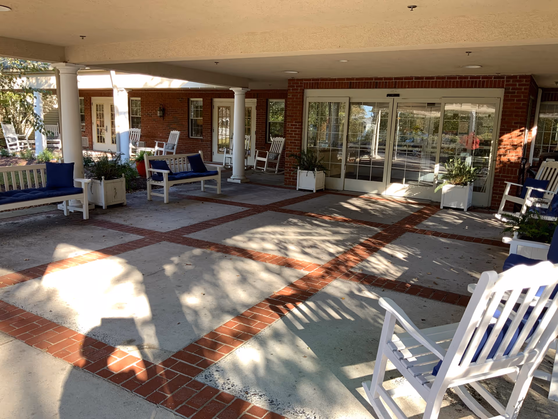 Covered outdoor seating area at Woodview Estates with white wooden benches and rocking chairs featuring blue cushions, surrounded by white columns and brick walls with glass doors leading inside.
