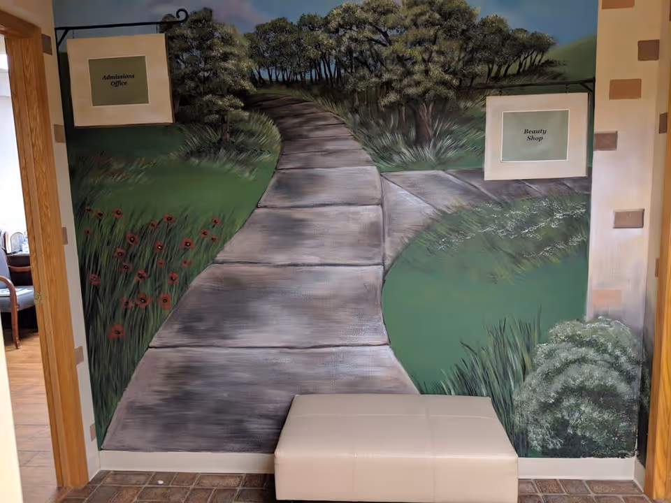 A painted mural on a wall depicting a pathway through a green landscape with trees and flowers. There are two signs on the mural, one on the left labeled 'Admissions Office' and one on the right labeled 'Beauty Shop'. In front of the mural is a white cushioned bench. The floor is tiled and a wooden door frame is partially visible on the left side.