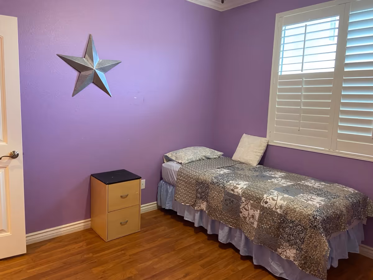 A small bedroom with purple walls featuring a single bed with a patterned quilt and two pillows. There is a wooden nightstand with two drawers next to the bed and a large metal star decoration on the wall. A window with white plantation shutters is on the right side, and the floor is wooden.