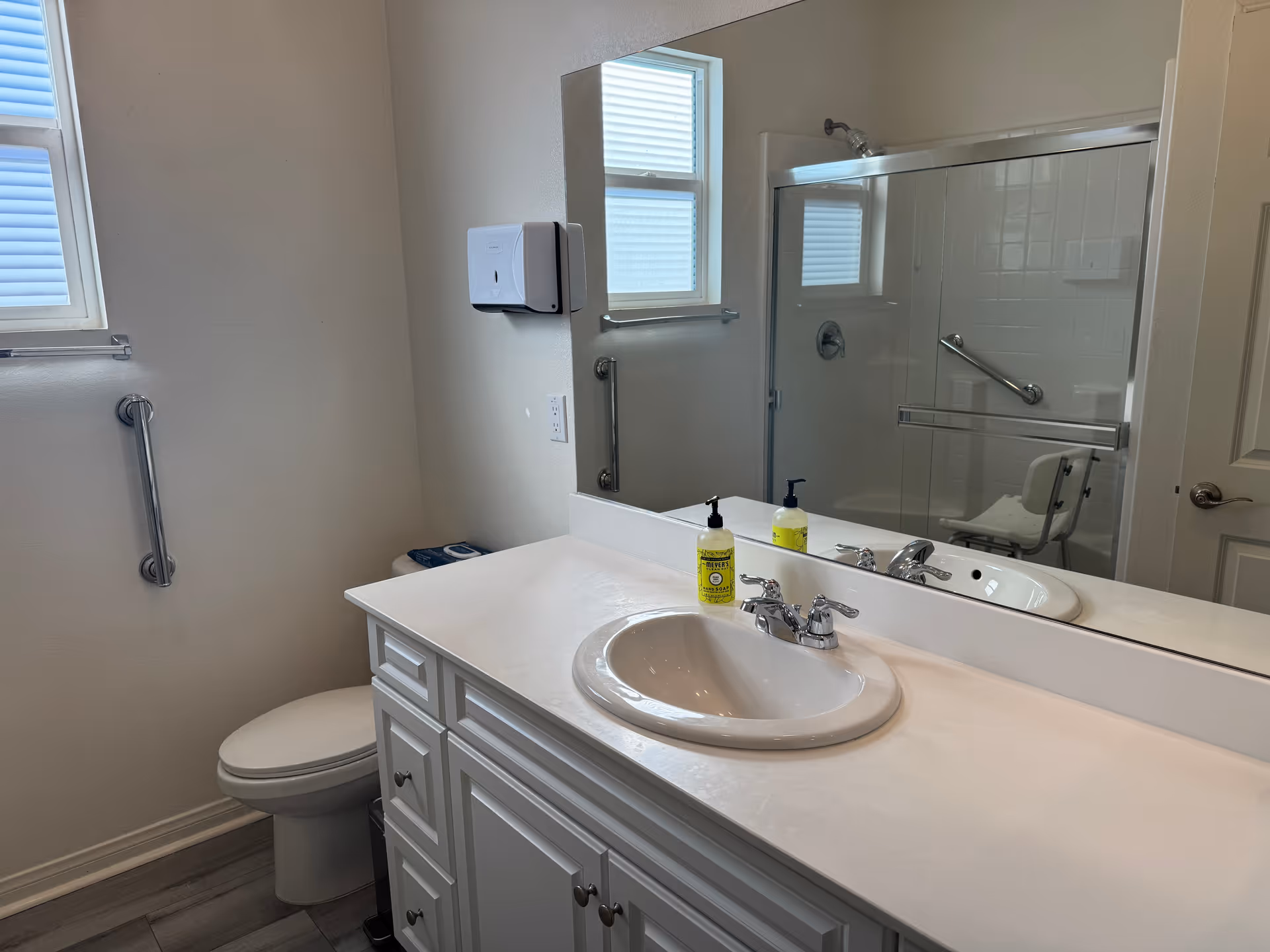 A clean and bright bathroom featuring a white sink with a chrome faucet on a white countertop, a large mirror above the sink, a toilet with a grab bar next to it, a window with blinds, and a glass-enclosed shower with a shower chair and grab bars inside.