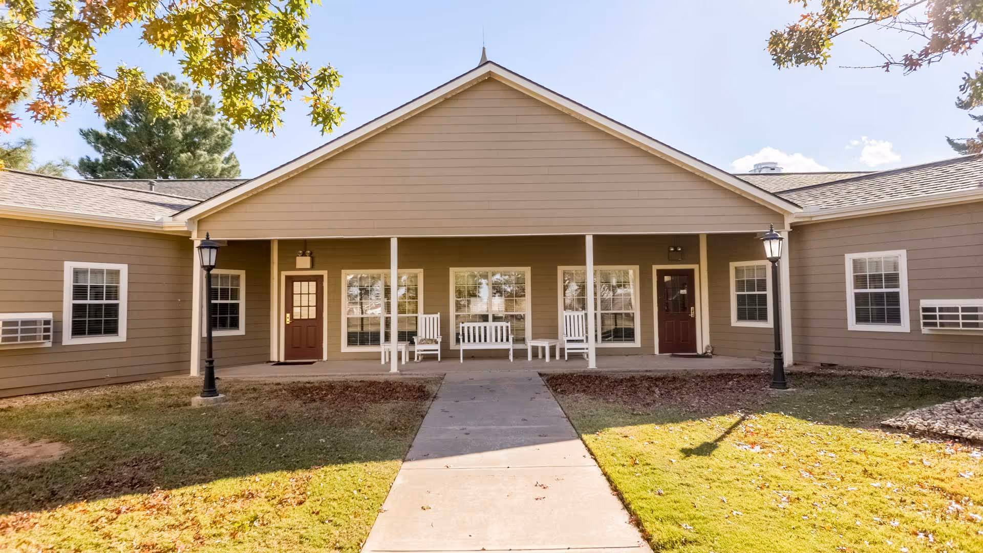 Exterior view of a single-story building with beige siding and a peaked roof. The building has two brown doors and several windows with white frames. There is a covered porch area with white rocking chairs and a bench. Two black lamp posts are positioned on either side of the concrete walkway leading to the porch. Trees with autumn-colored leaves frame the top of the image.