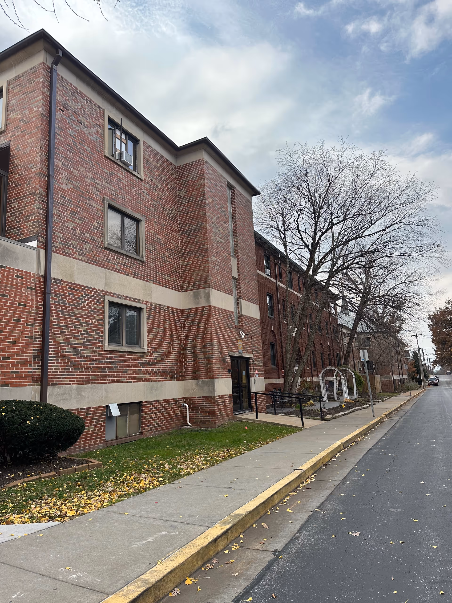 Exterior view of a multi-story brick building with several windows, a sidewalk, and a tree with no leaves. The sky is partly cloudy and there is a street running alongside the building.