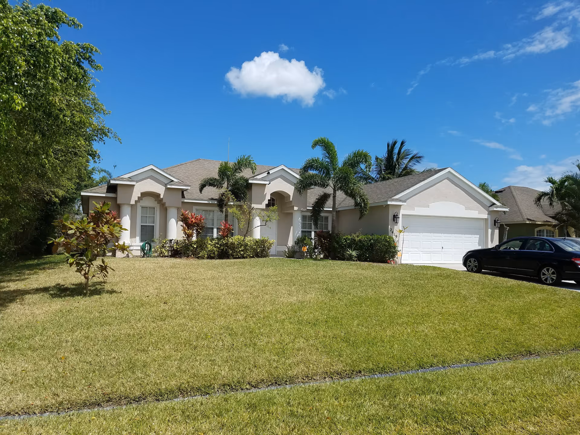 Single-story suburban house with a manicured front lawn, palm trees, and a car parked by the garage under a blue sky.
