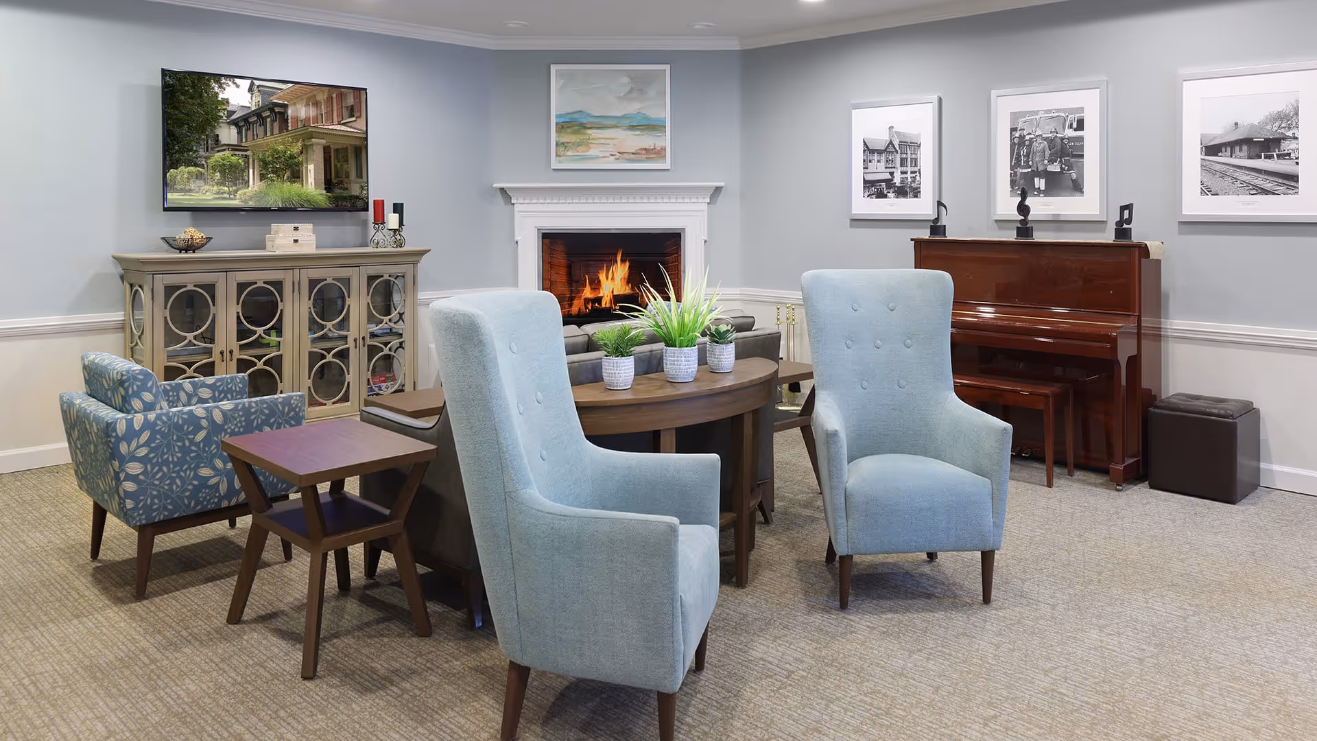 A cozy living room area with a lit fireplace, two light blue armchairs, a patterned armchair, a wooden side table, a round wooden table with potted plants, a wooden cabinet with a TV mounted above it, and a piano against the wall with framed black and white photographs hanging above.