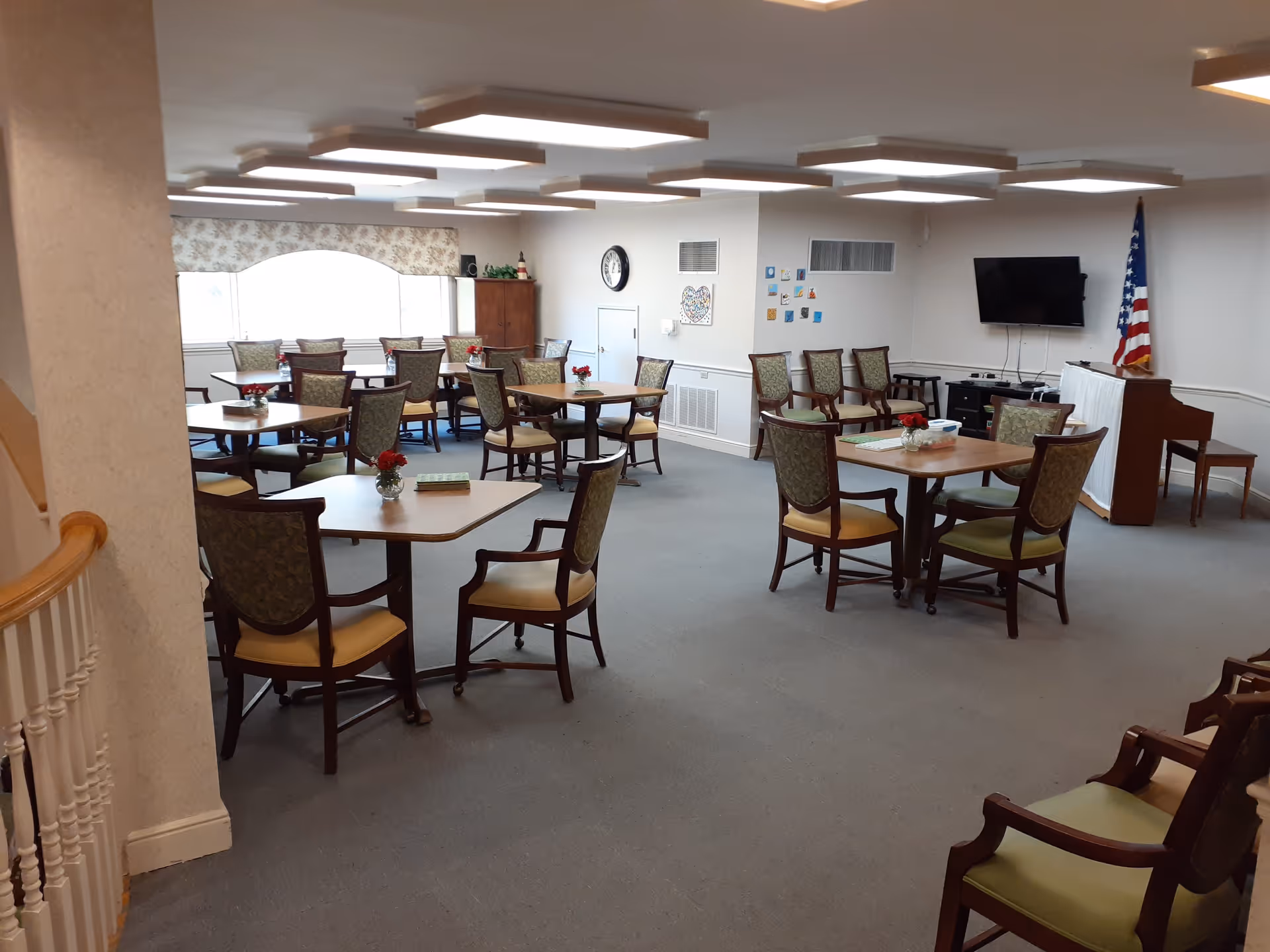 A spacious room with multiple square and rectangular tables surrounded by cushioned chairs. Each table has a small vase with red flowers. The room has a carpeted floor, a large window with floral curtains, a wall clock, a flat-screen TV mounted on the wall, an American flag, and a piano with a bench. The ceiling has modern rectangular light fixtures.
