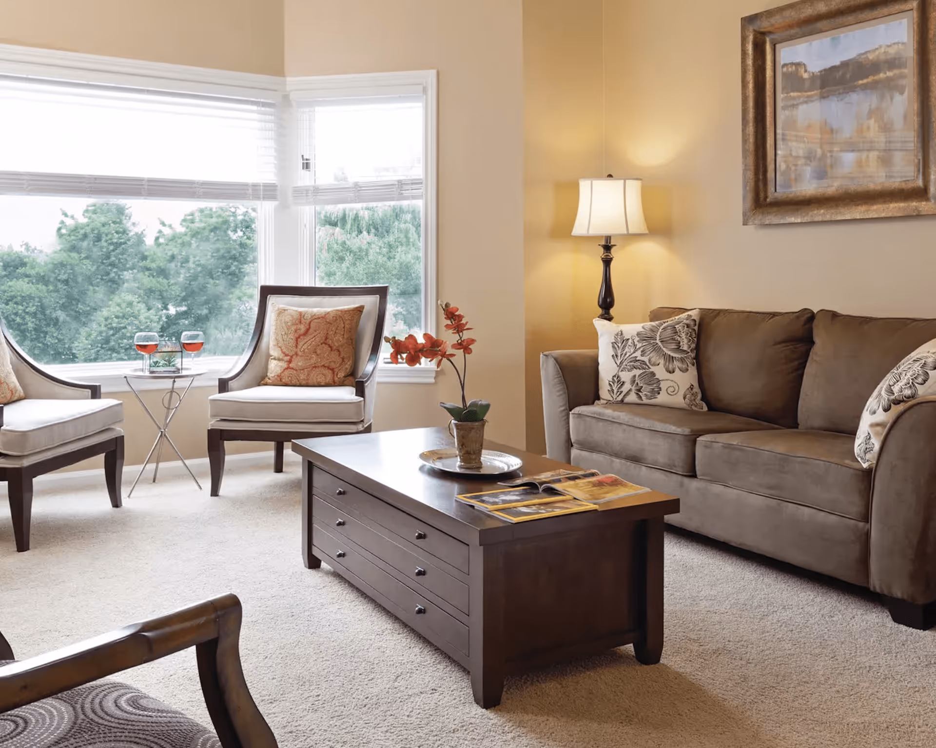 A cozy living room with a brown sofa adorned with floral pillows, two cushioned chairs with patterned pillows, a wooden coffee table with a potted plant and magazines, a floor lamp, and large windows showing green trees outside.