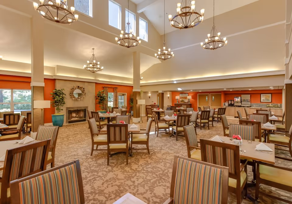 A spacious dining room with multiple wooden tables and striped cushioned chairs arranged neatly. The room features a high ceiling with several large chandeliers, a fireplace with a decorative mirror above it, large windows allowing natural light, and a beverage station in the back. The walls are painted in warm tones with orange accents.